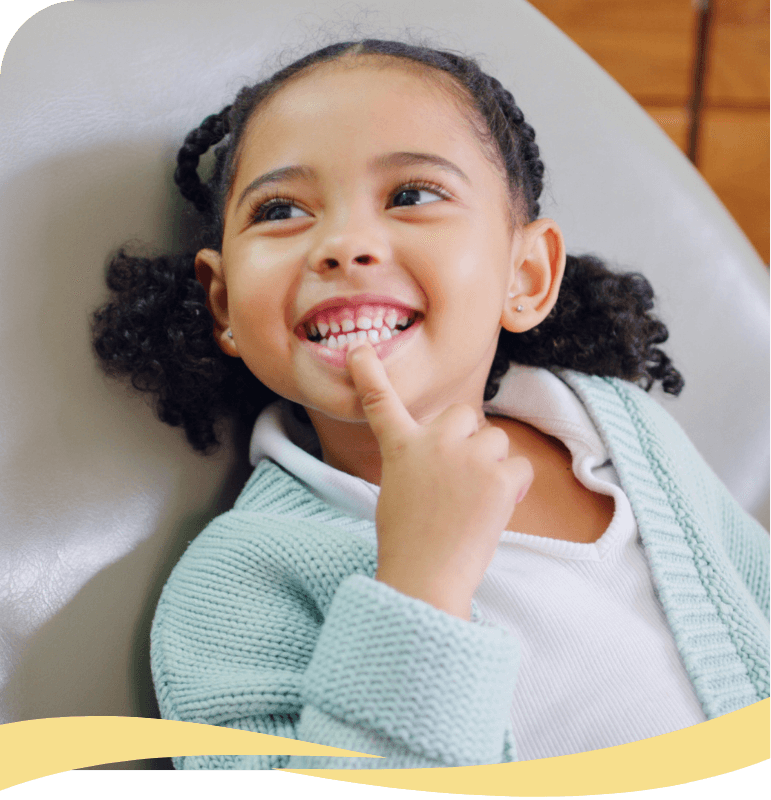 A young girl with curly hair in pigtails smiles brightly, showing her teeth, while sitting on a gray chair. She is wearing a light blue sweater and touching her mouth with her finger.