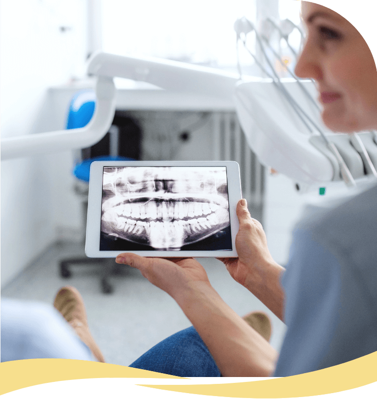 A person holds a tablet displaying a dental X-ray image in a dental office, while another person sits nearby in a dental chair. Dental equipment is visible in the background.