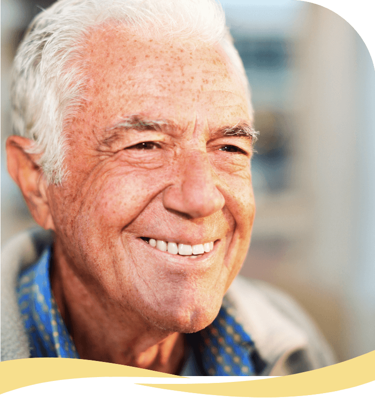 An older man with white hair smiles warmly, wearing a blue collared shirt and a jacket, with a softly blurred background and a yellow wave design at the bottom of the image.