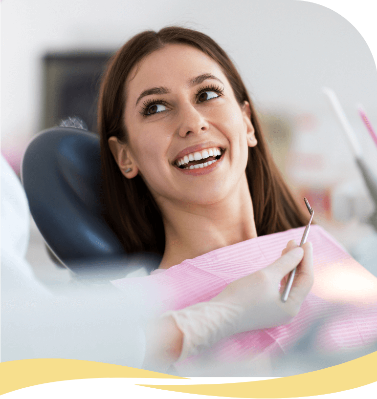 A young woman with long brown hair smiles while sitting in a dental chair, wearing a pink bib. A gloved dentist holds a dental tool near her. The atmosphere appears friendly and relaxed.