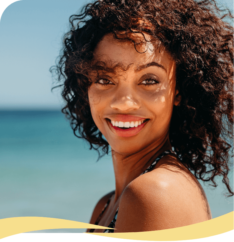 A woman with curly hair smiles brightly at the camera while standing on a sunny beach, with blue ocean and sky in the background.