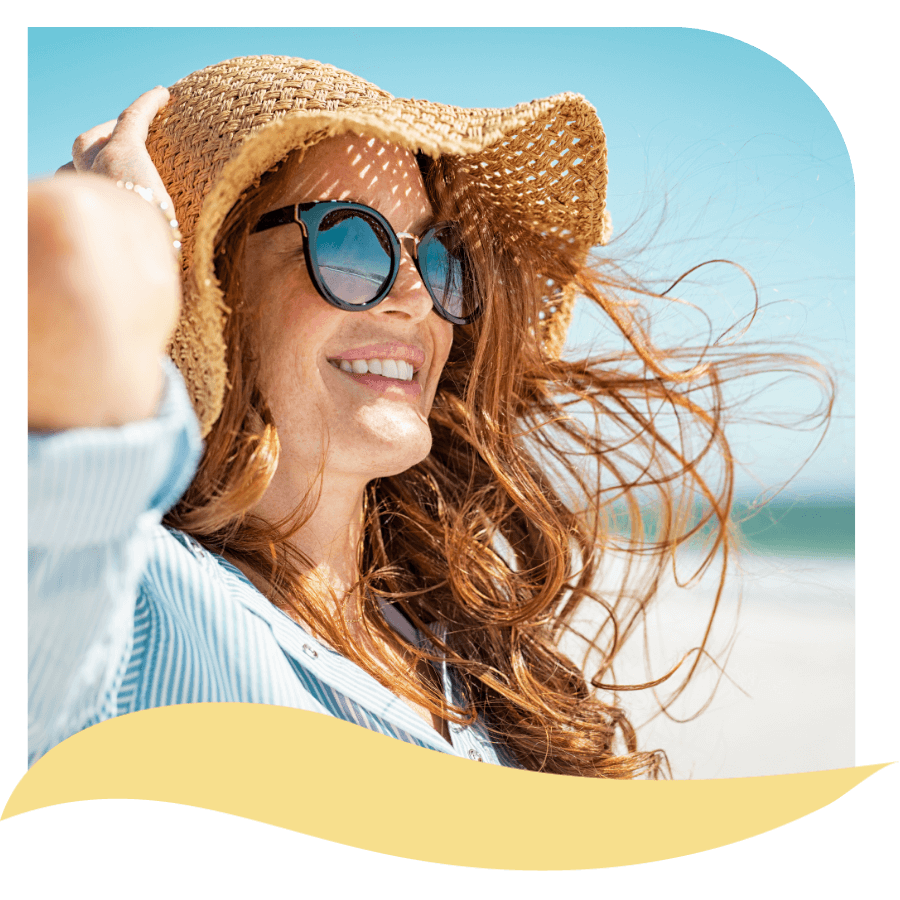 Smiling woman with long red hair wearing sunglasses and a straw hat stands on a sunny beach, with wind blowing her hair and a blue sky in the background.