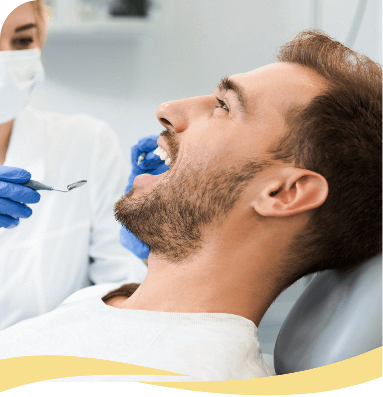 A man reclines in a dental chair with his mouth open while a dentist in gloves and a mask examines his teeth using dental tools.