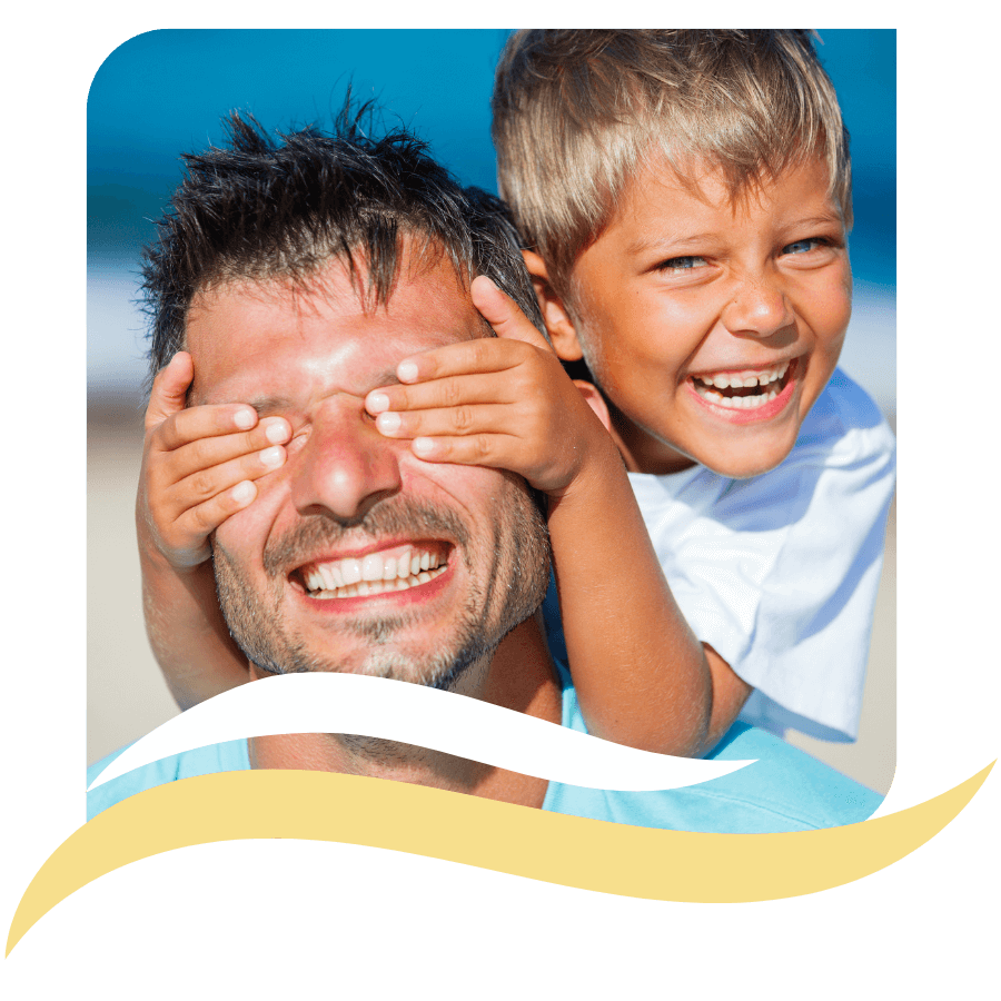 A smiling child playfully covers a mans eyes from behind while both laugh joyfully at the beach, with blue sky and ocean in the background.